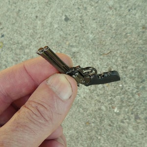 May include: A small, black metal replica of a revolver. The replica is detailed with a cylinder and a barrel.