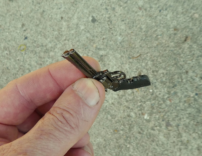 May include: A small, black metal replica of a revolver. The replica is detailed with a cylinder and a barrel.