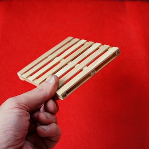 May include: A small wooden pallet made of thin strips of wood. The pallet is held in a hand against a red background.