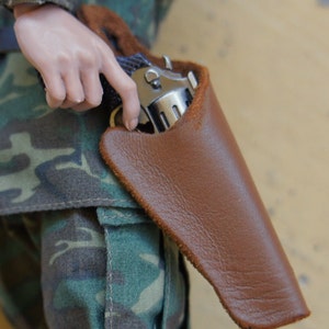 May include: A brown leather holster with a silver handgun inside. The holster is worn on a person's hip.