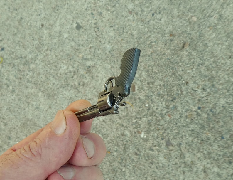 May include: A small, silver-coloured toy gun with a black handle. The gun is held in a person's hand.