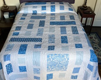 May include: A white and blue quilt with a geometric pattern covers a bed. The quilt features various shades of blue squares and rectangles, some with paisley and circular designs. A purple headboard is visible.