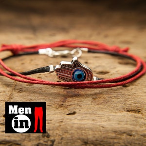 Könnte beinhalten: Ein rotes und schwarzes Armband aus Schnur mit einem silbernen Anhänger mit einem blauen bösen Auge. Der Anhänger hat die Form einer Hand mit einem Auge in der Handfläche.