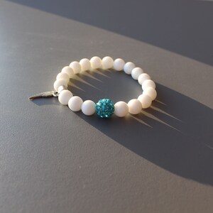 Op de afbeelding: Witte kralen armband met een turquoise kristallen bal bedel en een zilveren vleugel hanger.