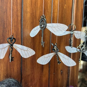 May include: A whimsical mobile with five antique-style keys, each with delicate wings, hanging from white strings against a wood background. The keys are adorned with various details, including a crown, a heart, and a rabbit.