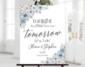 May include: A white wedding sign on a black easel, adorned with floral arrangements in shades of blue and cream. The sign reads "Tonight, We Celebrate with you Tomorrow, We say 'I do' Olivia & Stephen, August 18, 20XX."