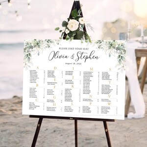 Puede incluir: Un cartel blanco de distribución de asientos para bodas con el texto Please Find Your Seat, Olivia & Stephen, y August 18, 202X. El cartel está decorado con hojas de acuarela verdes y se apoya en un caballete de madera oscura, mostrando los nombres.