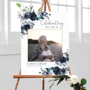 May include: A white memorial sign on a wooden easel, featuring a photo of a woman and floral arrangements in navy and white. The sign reads "Celebrating the life of" and includes the name "Karen Carter" and dates.