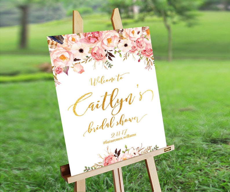 K&ouml;nnte beinhalten: Ein wei&szlig;es Willkommens-Schild mit goldenen Lettern f&uuml;r eine Brautparty. Das Schild zeigt ein Blumenmuster mit rosa und wei&szlig;en Blumen und gr&uuml;nen Bl&auml;ttern. Der Text auf dem Schild lautet "Willkommen zu Caitlyn's Bridal Shower 2.11.17 #futuremrs.williams".