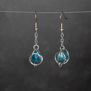 May include: A pair of handmade earrings with a silver wire cage design. Each earring features a teal-colored bead encased in the wire. One earring is teardrop-shaped, the other is spherical. The earrings hang from silver-colored ear wires.