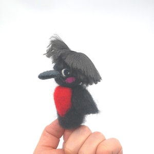 May include: A black and red felt bird with a black felt beak and a black felt crest. The bird is perched on a finger.