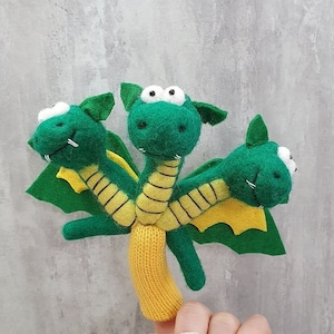 May include: A green three-headed finger puppet dragon with yellow stripes and wings. The dragon has white eyes and a yellow knitted body.
