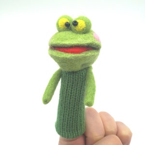 Puede incluir: Títere de dedo de fieltro verde de una rana con ojos amarillos, boca roja y mejillas rosadas. La rana lleva un suéter de punto verde.