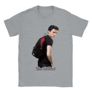 Puede incluir: Camiseta gris con un gráfico de Tom Holland y una mochila negra. La mochila tiene un interior rojo. El texto "Tom Holland" está impreso en cursiva en la parte inferior de la imagen.