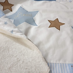 Könnte beinhalten: Eine weiße Babydecke mit einem Muster aus blauen und braunen Sternen. Die Decke hat eine weiche, flauschige Textur und ist aus einem Patchwork aus verschiedenen Stoffen gefertigt. Die Sterne sind mit dem Namen "Linus" bestickt.