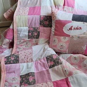 Könnte beinhalten: Eine rosafarbene und weiße Patchwork-Decke mit zwei Kissen. Die Kissen sind ebenfalls aus Patchwork und haben die Namen "Norya" und "Assia" gestickt.