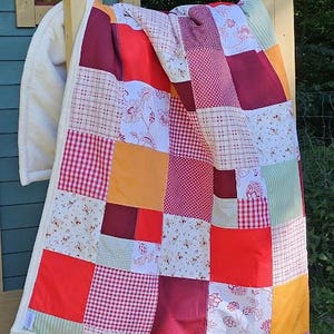 Könnte beinhalten: Ein Patchwork-Quilt mit einer Vielzahl von rot-, orange- und weiß gemusterten Quadraten. Der Quilt ist über einer Holzkonstruktion drapiert und zeigt eine Mischung aus floralen, Vichy- und unifarbenen Stoffdesigns. Der Rand des Quilts ist cremefarben.