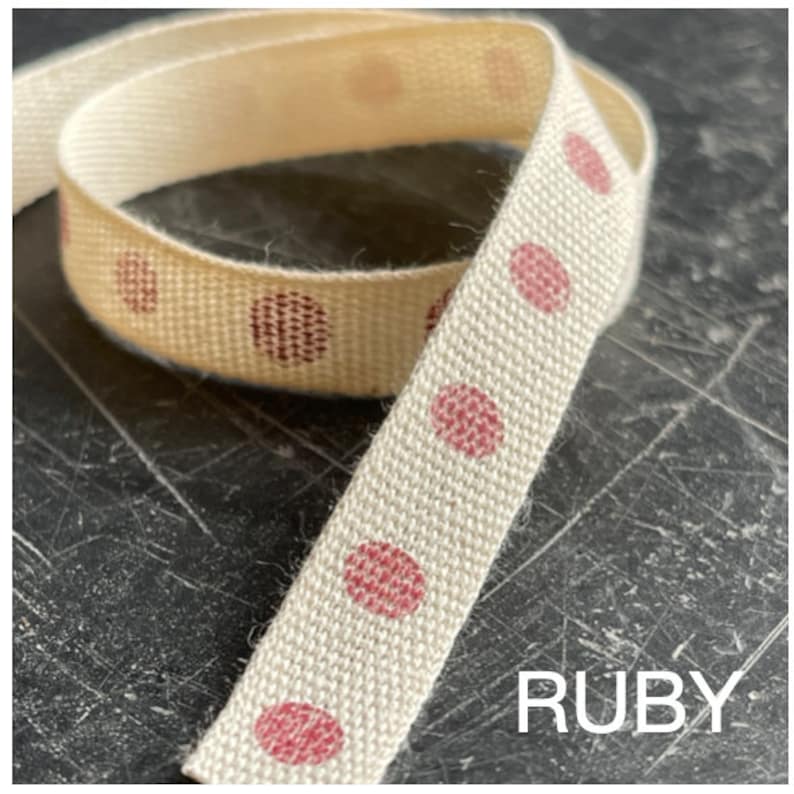 May include: A close-up of a white cotton ribbon with pink polka dots. The ribbon is folded in half and the word "RUBY" is printed in white on a black background.