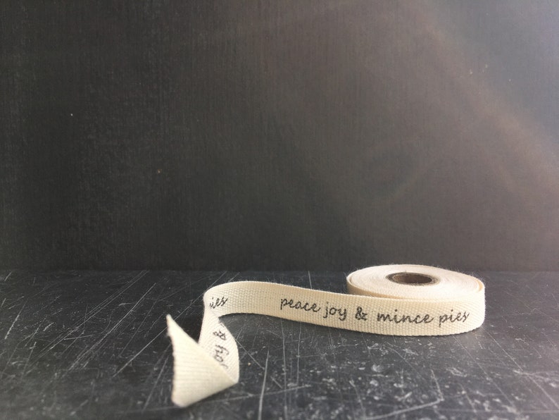 May include: A roll of natural coloured fabric ribbon with the text "peace joy & mince pies" printed in black ink.