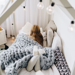 May include: A young person is curled up in a bed with a chunky knit blanket, reading a book. The bed is decorated with pillows and fairy lights.