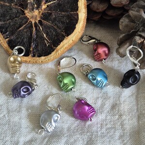 Peut inclure: Neuf marqueurs de points en forme de crâne dans différentes couleurs, notamment argent, violet, vert, bleu, rose et rouge. Les marqueurs sont sur un fond de tissu blanc.