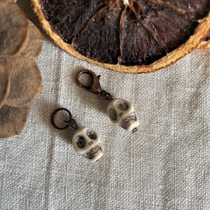 Peut inclure: Deux petits breloques en forme de crâne blanc cassé avec des orbites sombres et un fermoir et un anneau de couleur bronze. Les breloques sont posées sur un fond en lin neutre, avec une tranche d'orange séchée et des gousses en arrière-plan.