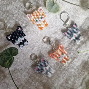 May include: Several beaded cat keyrings in different colours, including black, orange, and grey. Each keyring features a cat design with a silver-coloured clasp or ring for attachment. The cats are made of small, colourful beads.