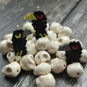May include: Three beaded raven pendants with yellow beaks and red eyes rest on a bed of white skull beads. The ravens are black with yellow and red accents. The skull beads have a marbled appearance.