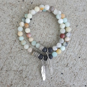 May include: A double-strand bracelet made with light green, brown, and white beads. The bracelet has two silver charms and two black lava beads with silver feather charms.