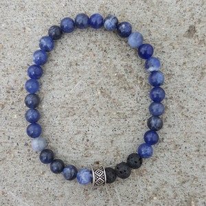 May include: A beaded bracelet featuring blue sodalite and black lava rock beads. The bracelet is strung on an elastic band and includes a silver-colored spacer bead with a geometric design. The beads are round and vary in shades of blue.