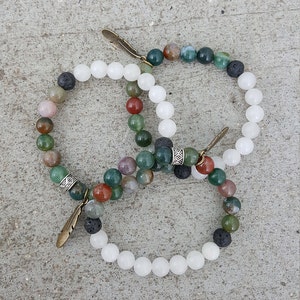 May include: Two bracelets with a mix of green, brown, white, and black beads. One bracelet has white beads and the other has a mix of colors. Both bracelets have a small feather charm.