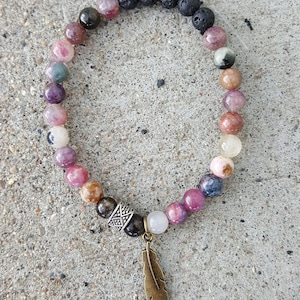 May include: A multi-colored gemstone bracelet with a feather charm. The bracelet features black lava beads, pink and green gemstone beads, and a silver bead with a decorative design. The feather charm is made of bronze.