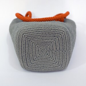 May include: A gray crocheted tote bag with orange rope handles. The bag is shown from the bottom, revealing the crocheted pattern.