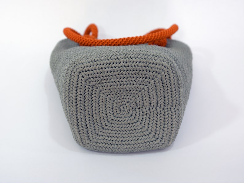 May include: A gray crocheted tote bag with orange rope handles. The bag is shown from the bottom, revealing the crocheted pattern.