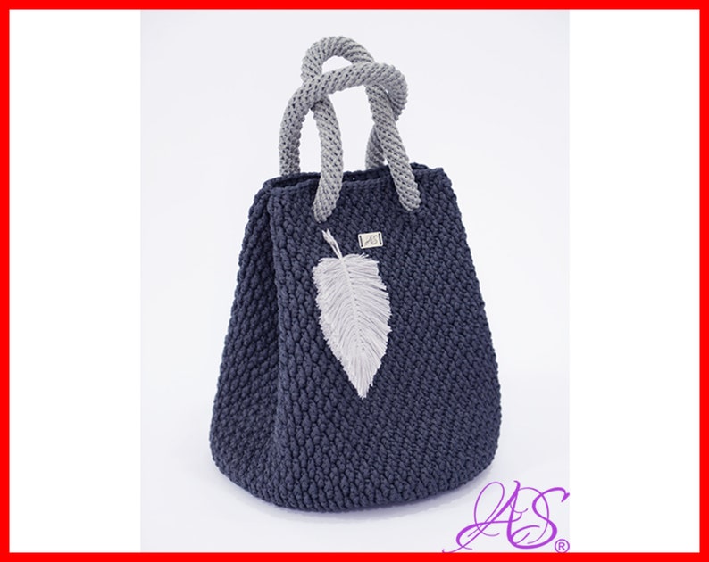 May include: A dark blue crocheted tote bag with a gray crocheted handle and a gray feather charm. The bag has a textured surface and is made of thick yarn.
