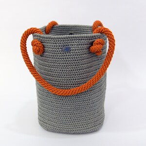 May include: A gray crocheted tote bag with orange rope handles. The bag has a woven texture and is made with a thick yarn.