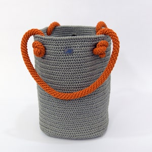 May include: A gray crocheted tote bag with orange rope handles. The bag has a woven texture and is made with a thick yarn.