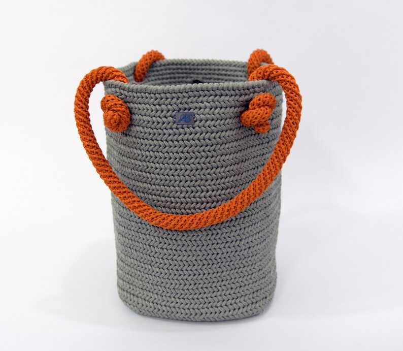 May include: A gray crocheted tote bag with orange rope handles. The bag has a woven texture and is made with a thick yarn.