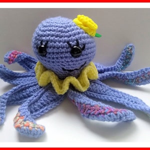 May include: A crocheted purple octopus with yellow and green accents. The octopus has a yellow flower on its head and a yellow ruffle around its neck. The octopus has eight tentacles with colorful stripes.