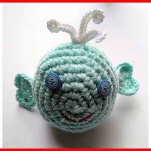 May include: A crocheted whale toy with blue eyes and a red mouth. The whale is light blue and has white crocheted fins and a white crocheted spout.