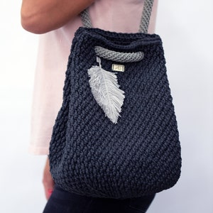 May include: A dark gray crocheted tote bag with a gray rope handle and a white feather charm. The bag has a small label with the text "AS" on it.