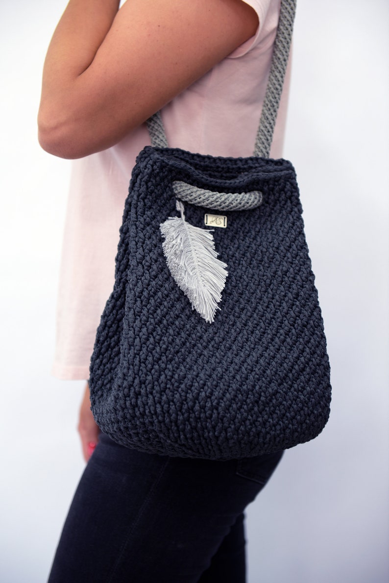 May include: A dark gray crocheted tote bag with a gray rope handle and a white feather charm. The bag has a small label with the text "AS" on it.
