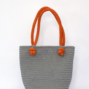 May include: A gray crocheted tote bag with orange rope handles. The bag has a knotted detail on each handle.