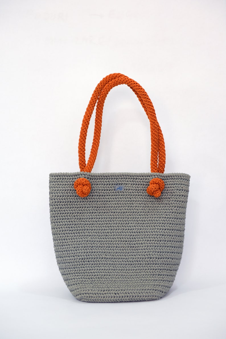 May include: A gray crocheted tote bag with orange rope handles. The bag has a knotted detail on each handle.