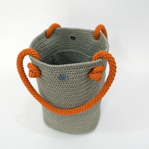 May include: A gray crocheted tote bag with orange rope handles. The bag has a small blue label with the text "JBS" on it.