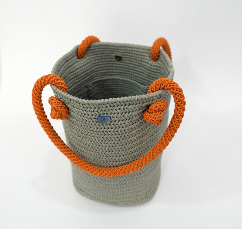 May include: A gray crocheted tote bag with orange rope handles. The bag has a small blue label with the text "JBS" on it.