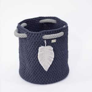 May include: A dark blue crocheted storage basket with gray rope handles and a white feather accent. The basket is made of a thick, textured yarn and has a round shape.