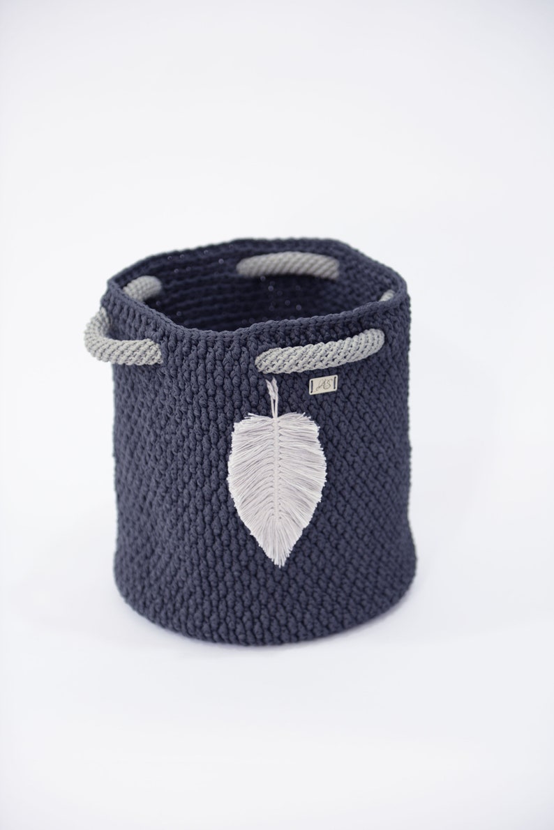 May include: A dark blue crocheted storage basket with gray rope handles and a white feather accent. The basket is made of a thick, textured yarn and has a round shape.