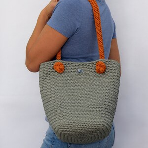 May include: A gray crocheted tote bag with orange handles and pom-poms. The bag has a small blue label with the letters "AS" on it.