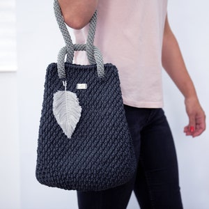 May include: A dark gray crocheted tote bag with a gray rope handle and a feather charm. The bag has a small silver tag with the letters "MC" on it.
